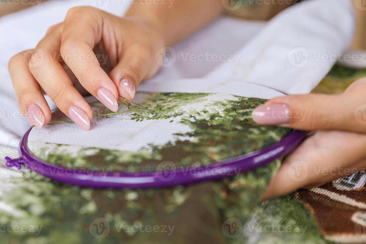 A girl cross-stitching. Close-up of hands carefully maneuvering a needle through the fabric. A purple embroidery hoop holds the fabric. Close-up of hands with a needle threading the fabric. photo