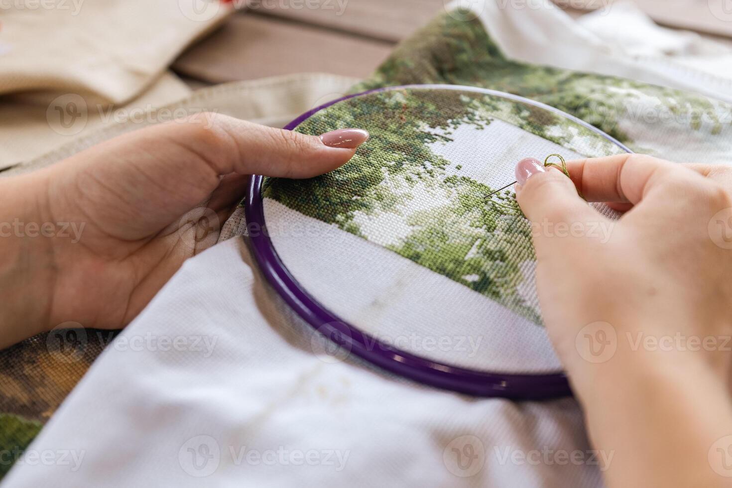 A girl cross-stitching. Close-up of hands carefully maneuvering a needle through the fabric. A purple embroidery hoop holds the fabric. Close-up of hands with a needle threading the fabric. photo