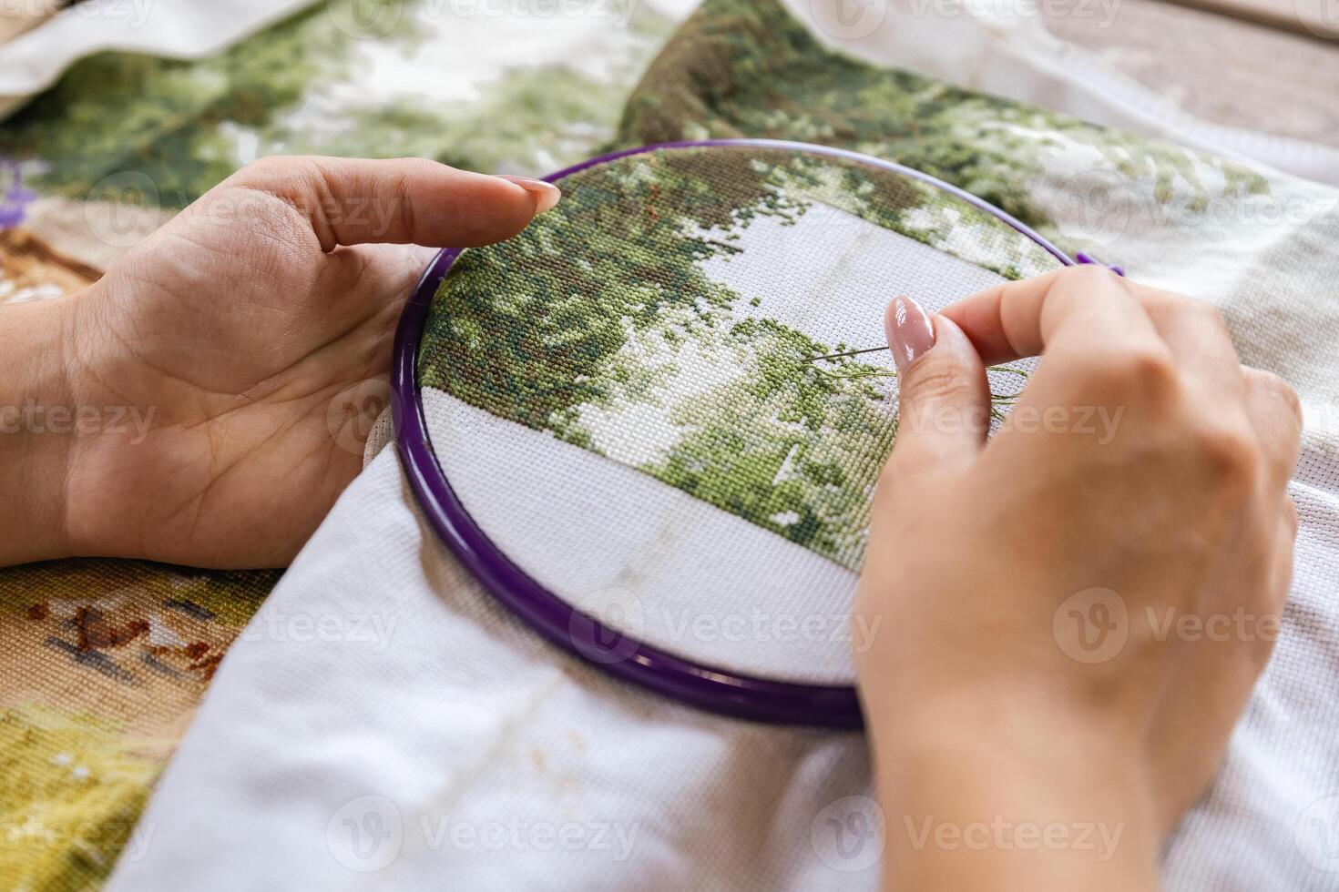 A girl cross-stitching. Close-up of hands carefully maneuvering a needle through the fabric. A purple embroidery hoop holds the fabric. Close-up of hands with a needle threading the fabric. photo