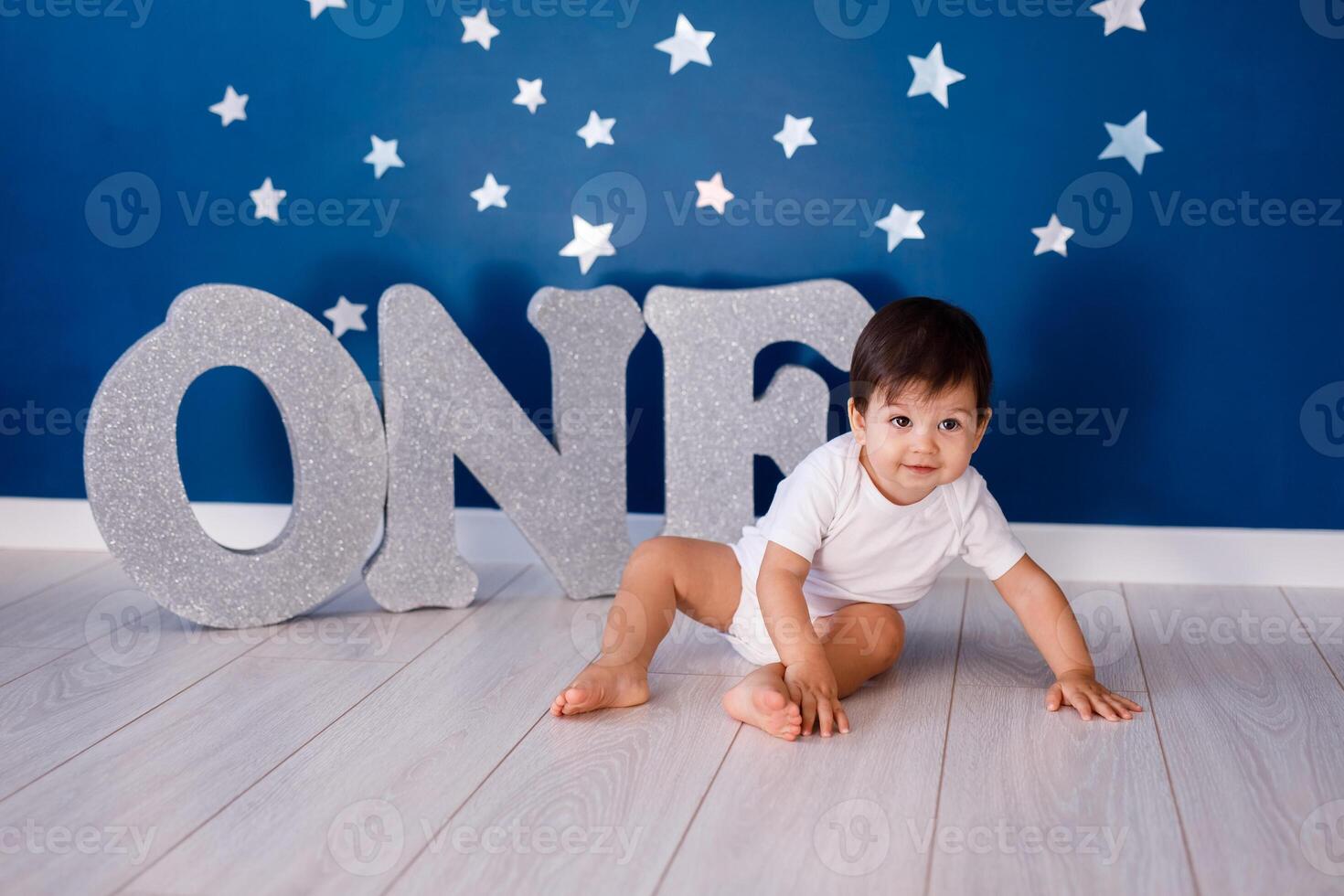 One year old baby boy in white t-shirt celebrates first birthday near silver letters ONE on blue background with stars photo