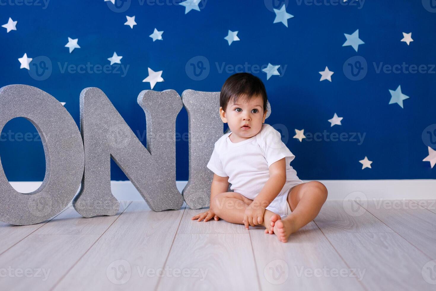 One year old baby boy in white t-shirt celebrates first birthday near silver letters ONE on blue background with stars photo