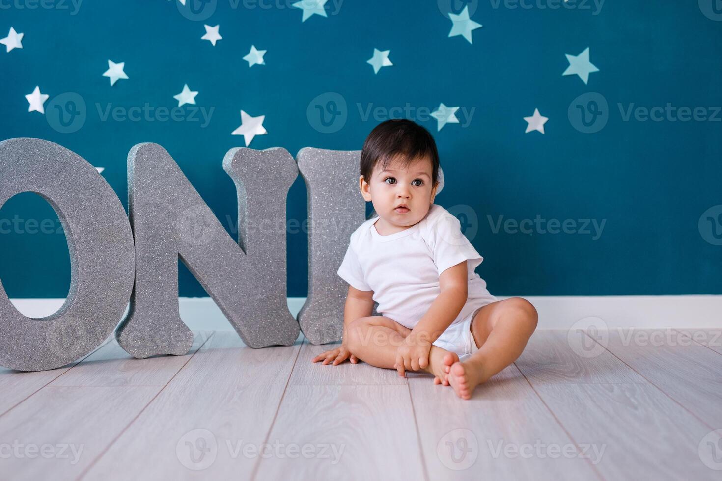 One year old baby boy celebrates birthday near silver letters ONE on blue background with stars photo