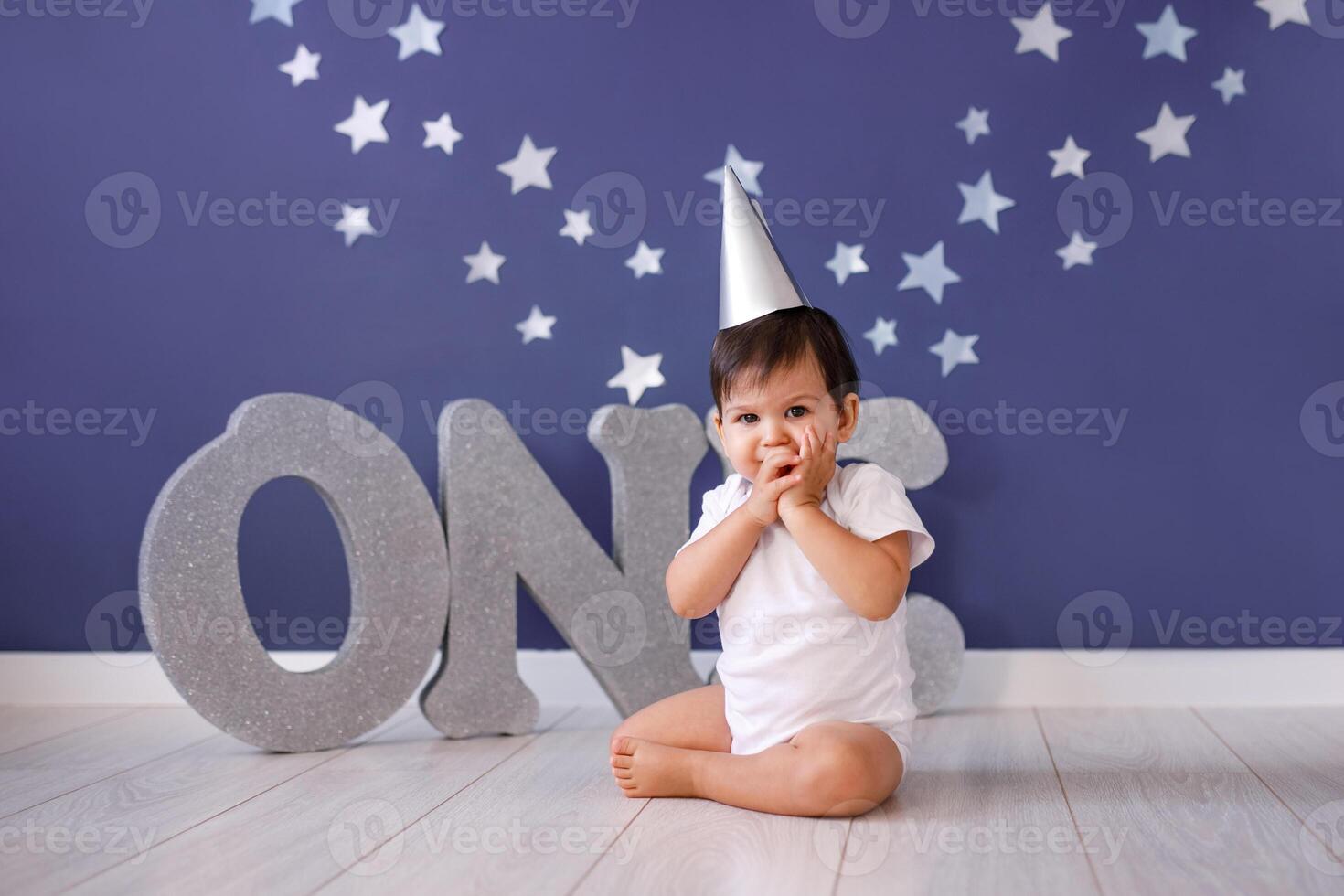 One year old baby boy celebrates birthday near silver letters ONE on blue background with stars photo