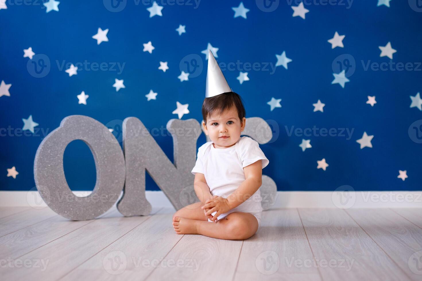 One year old baby boy celebrates birthday near silver letters ONE on blue background with stars photo