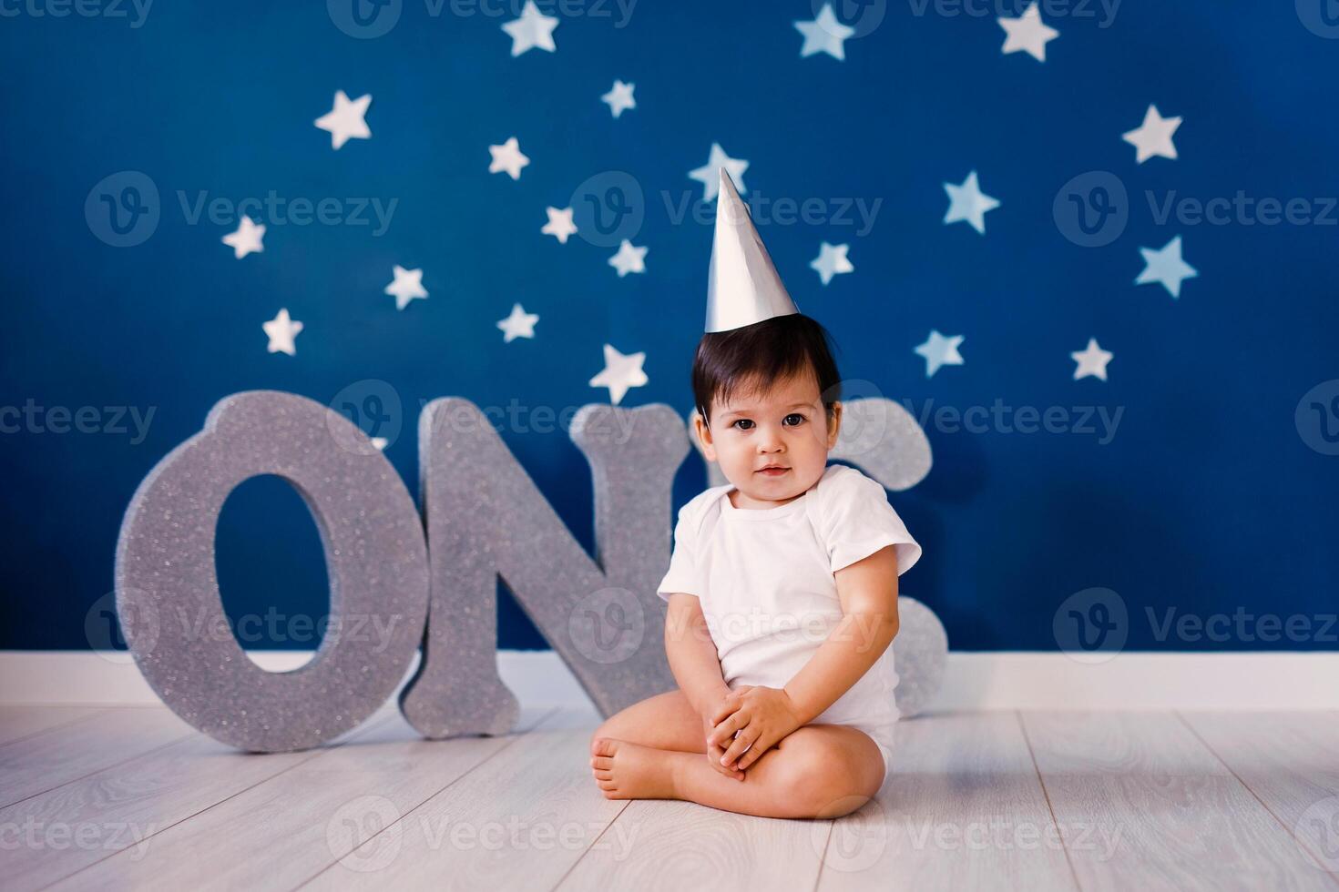 Baby boy of one year old sits on the floor on a blue background with stars and big silver letters one photo