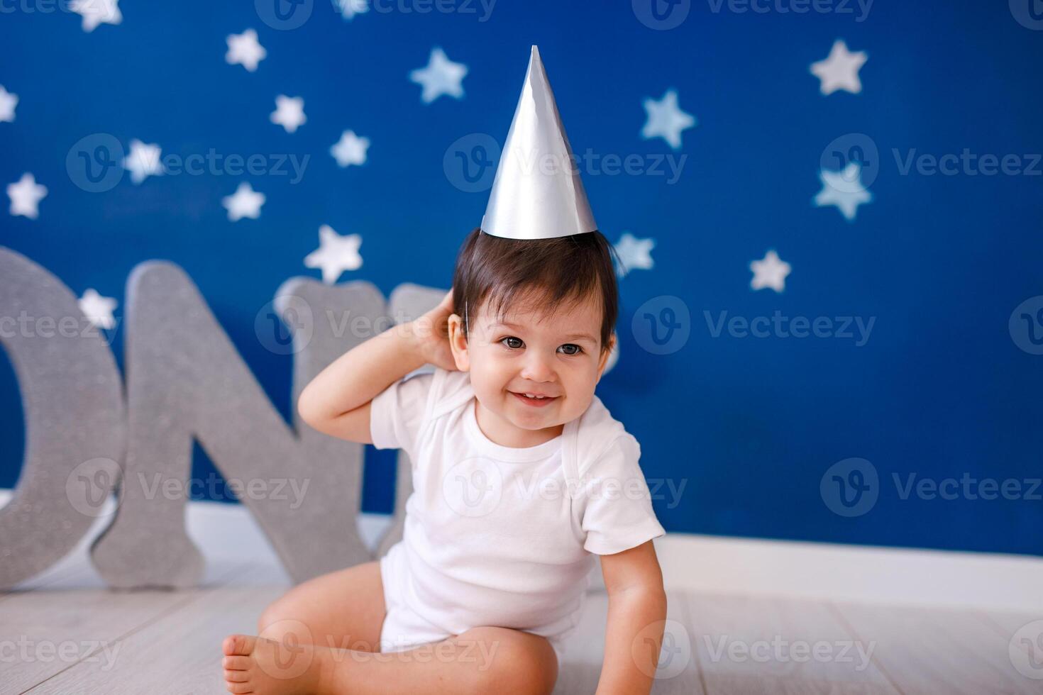 One year old baby boy celebrates birthday near silver letters ONE on blue background with stars photo