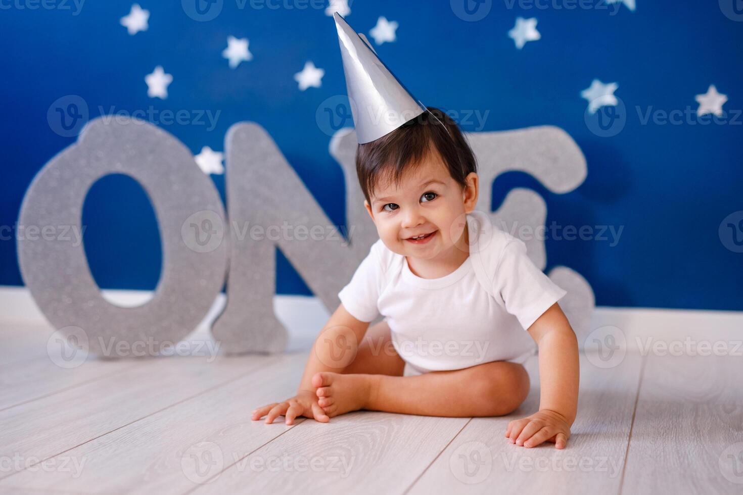 One year old baby boy celebrates birthday near silver letters ONE on blue background with stars photo