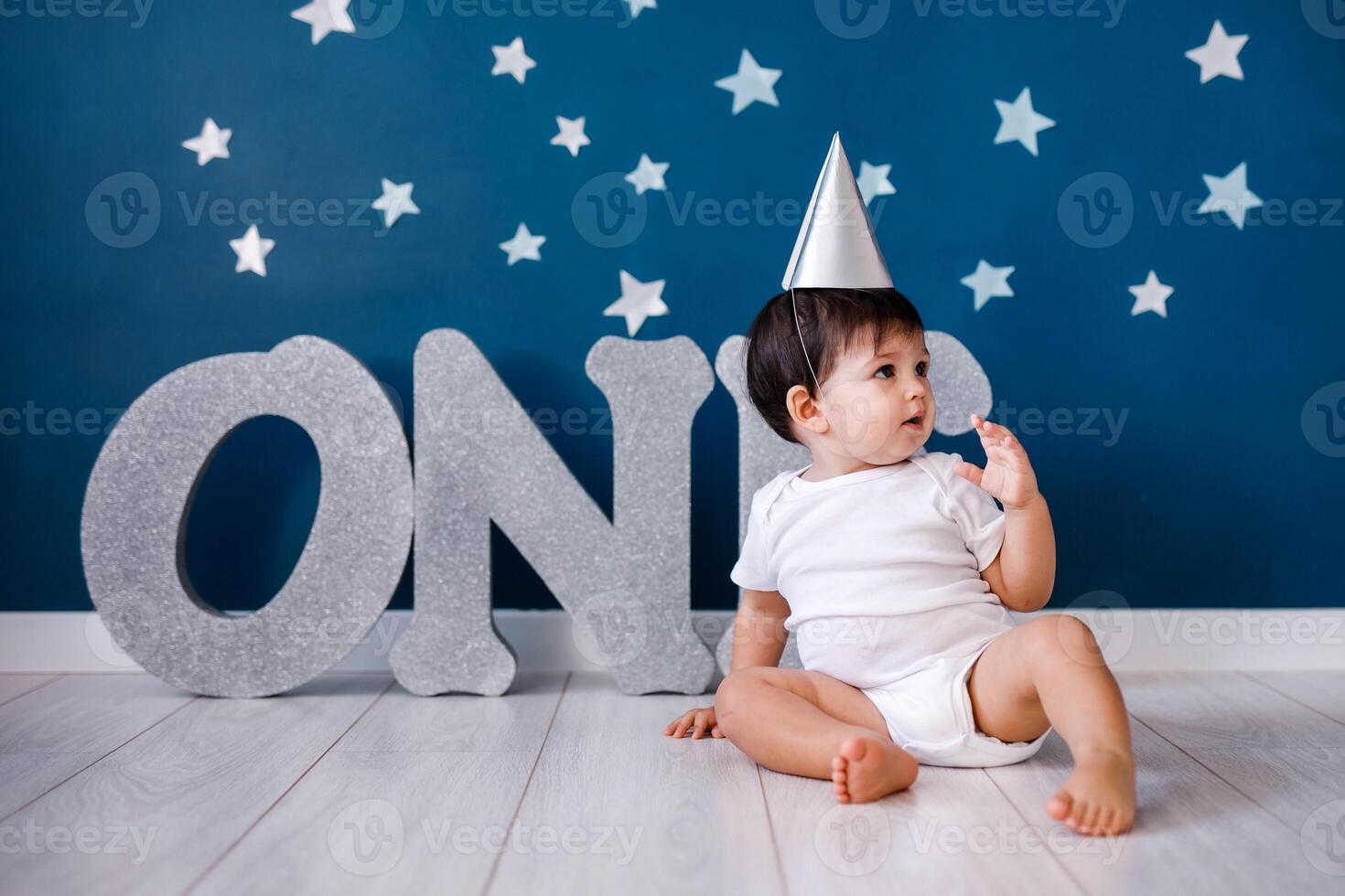 Baby boy of one year old wearing a white body and a festive paper hat sits on the floor on a blue background photo