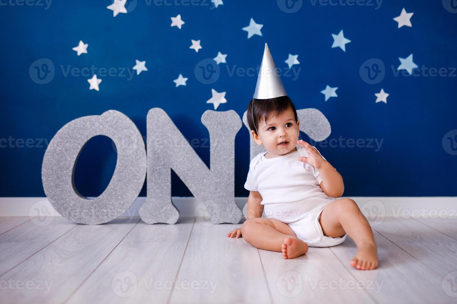 Baby boy of one year old wearing a white body and a festive paper hat sits on the floor photo