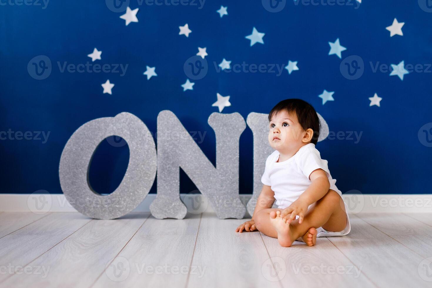 One year old baby boy in white t-shirt celebrates first birthday near silver letters ONE on blue background with stars photo