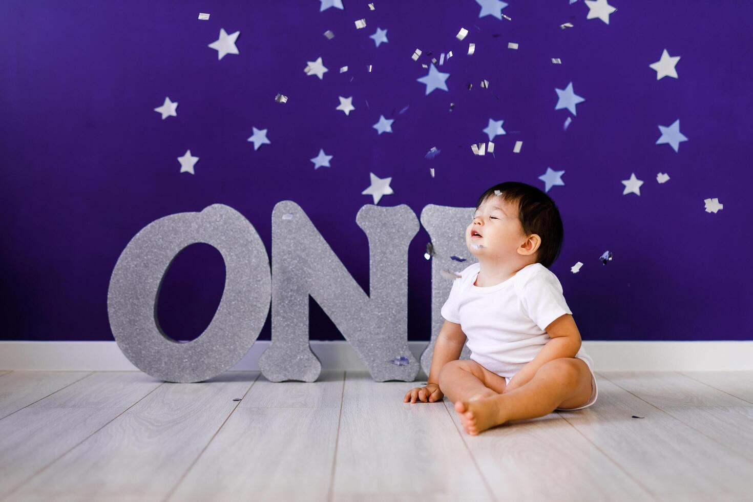 One year old baby boy celebrates birthday near silver letters ONE on blue background with stars photo