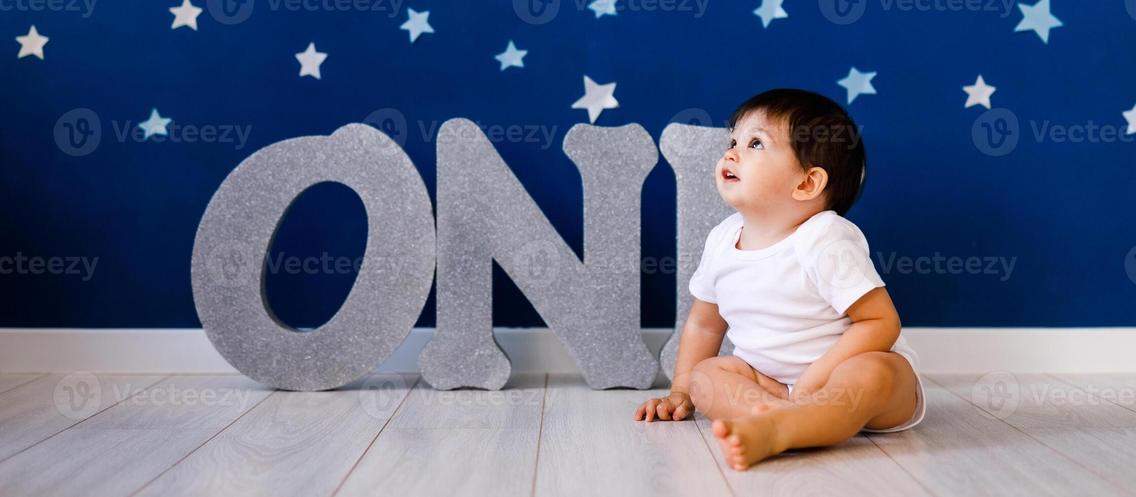 One year old baby boy celebrates birthday near silver letters ONE on blue background with stars photo