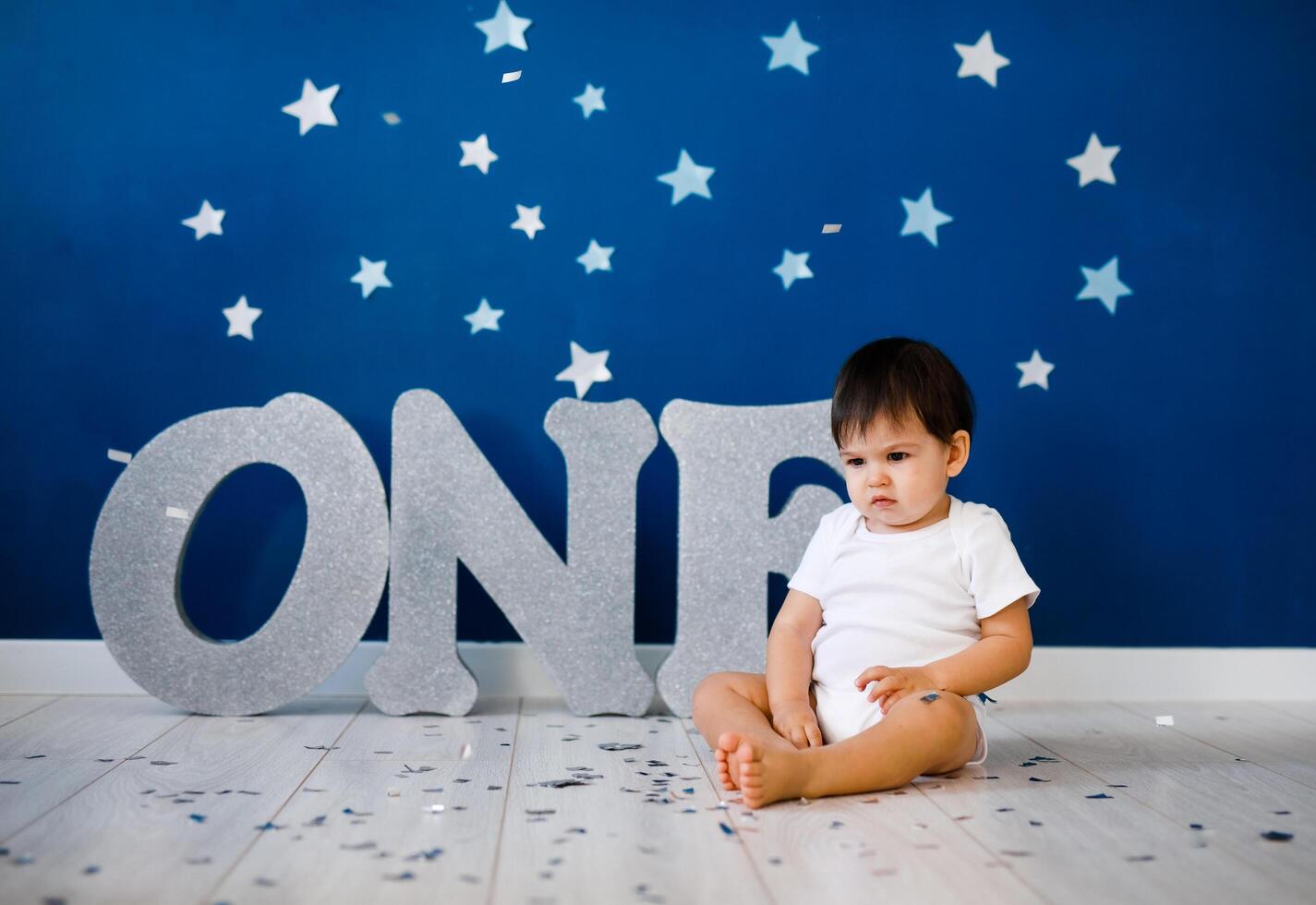 One year old baby boy celebrates birthday near silver letters ONE on blue background with stars photo