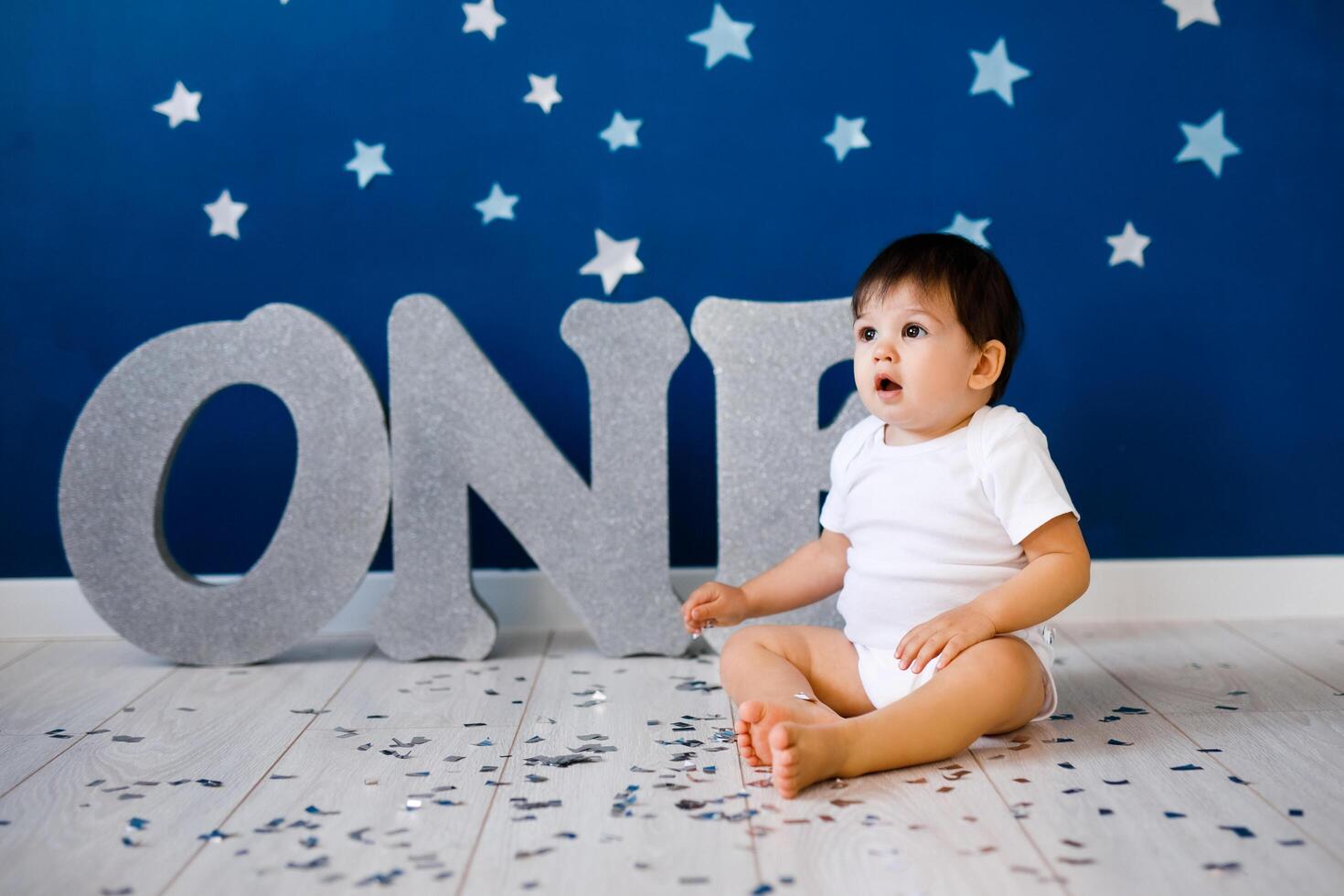 One year old baby boy in white t-shirt celebrates first birthday near silver letters ONE on blue background with stars photo