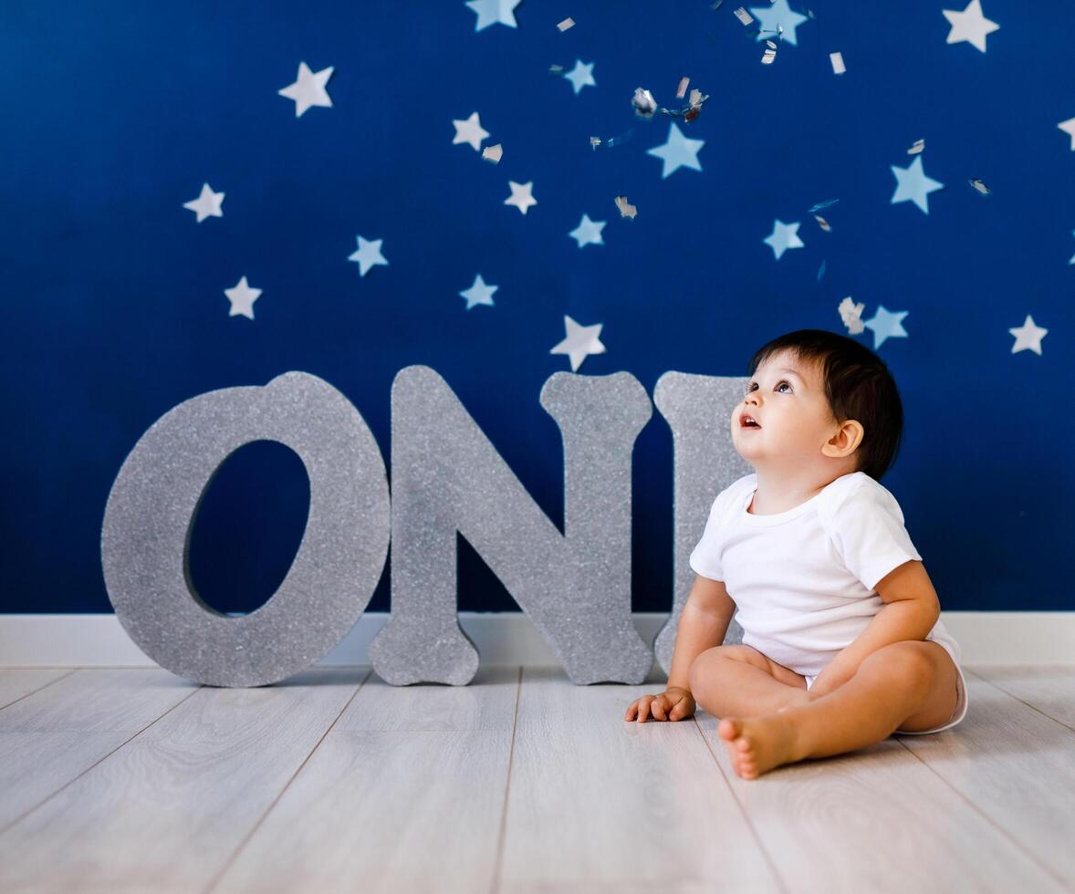 One year old baby boy celebrates birthday near silver letters ONE on blue background with stars photo