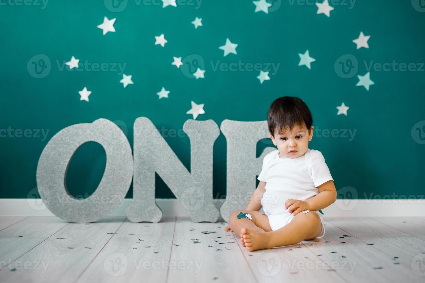 One year old baby boy celebrates birthday near silver letters ONE on blue background with stars photo