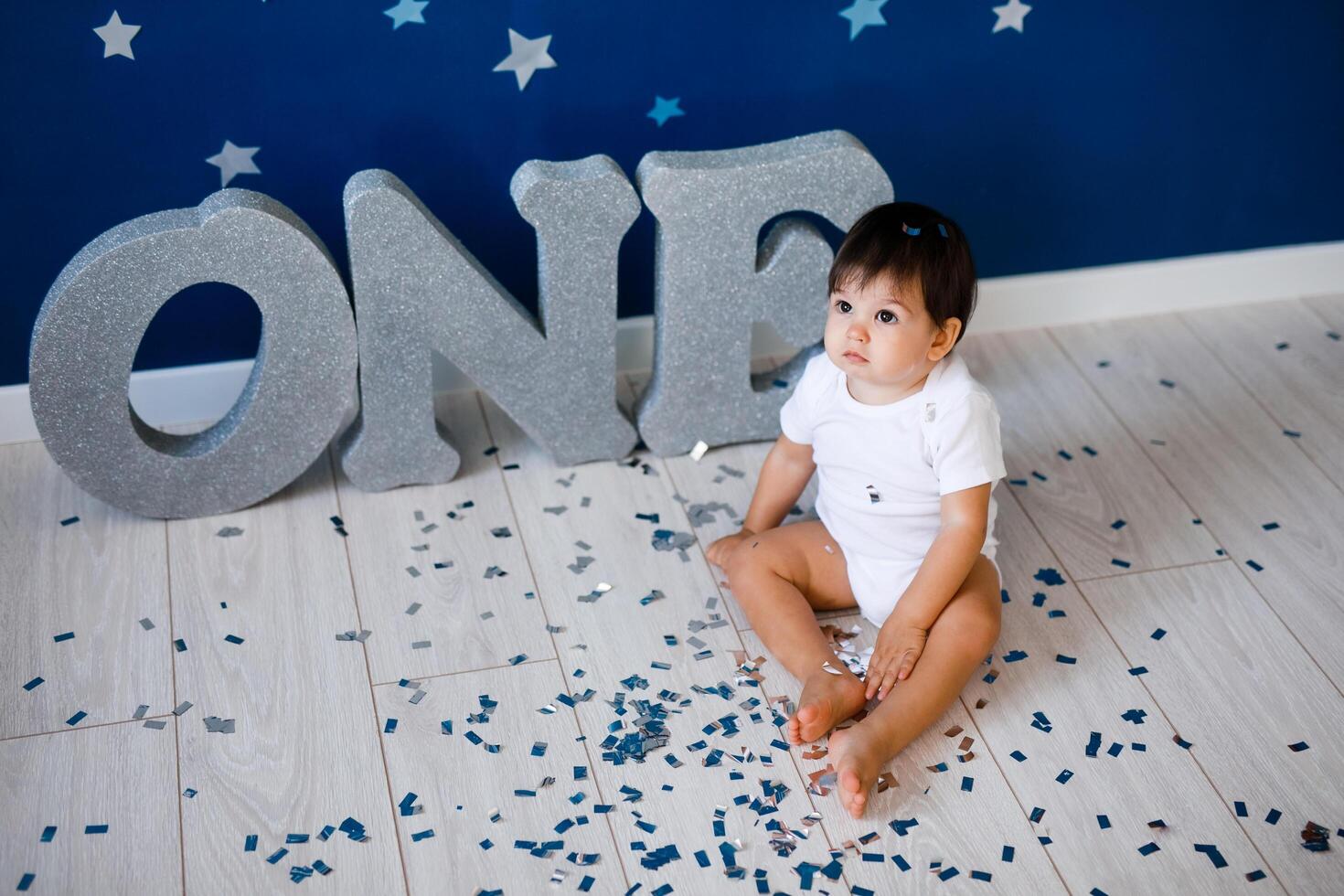 One year old baby boy celebrates birthday near silver letters ONE on blue background with stars photo