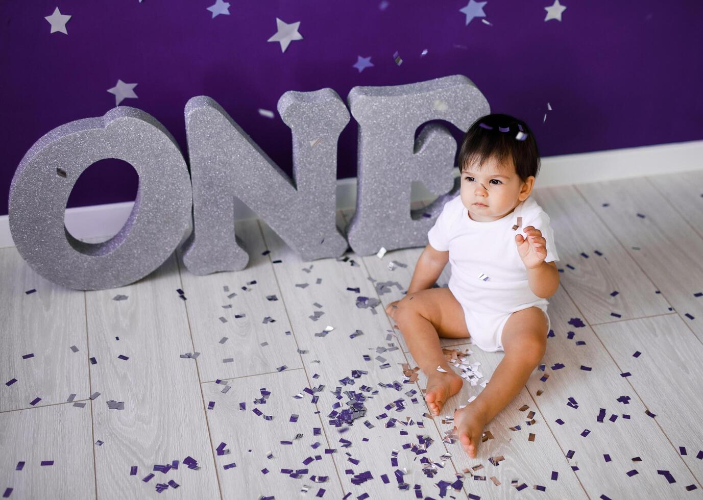 One year old baby boy in white t-shirt celebrates first birthday near silver letters ONE on purple background with stars photo