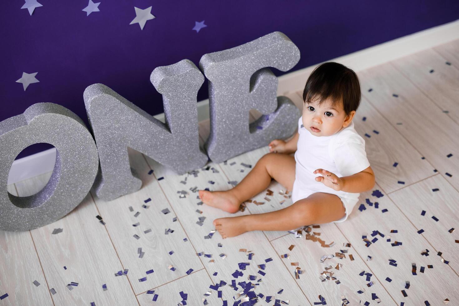 One year old baby boy celebrates birthday near silver letters ONE on blue background with stars photo