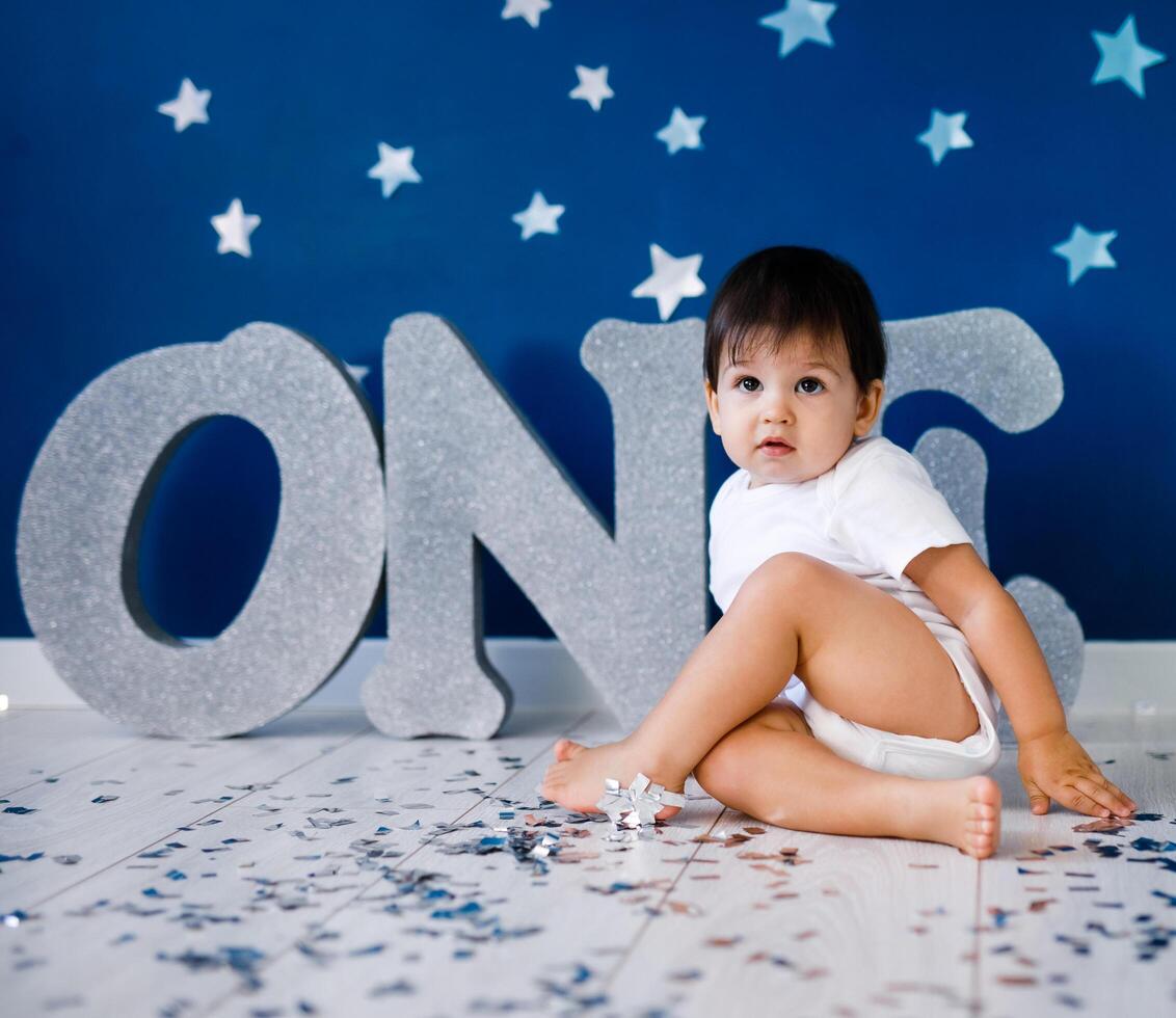 One year old baby boy in white t-shirt celebrates birthday near silver letters ONE on blue background with stars and silver sparkles. photo