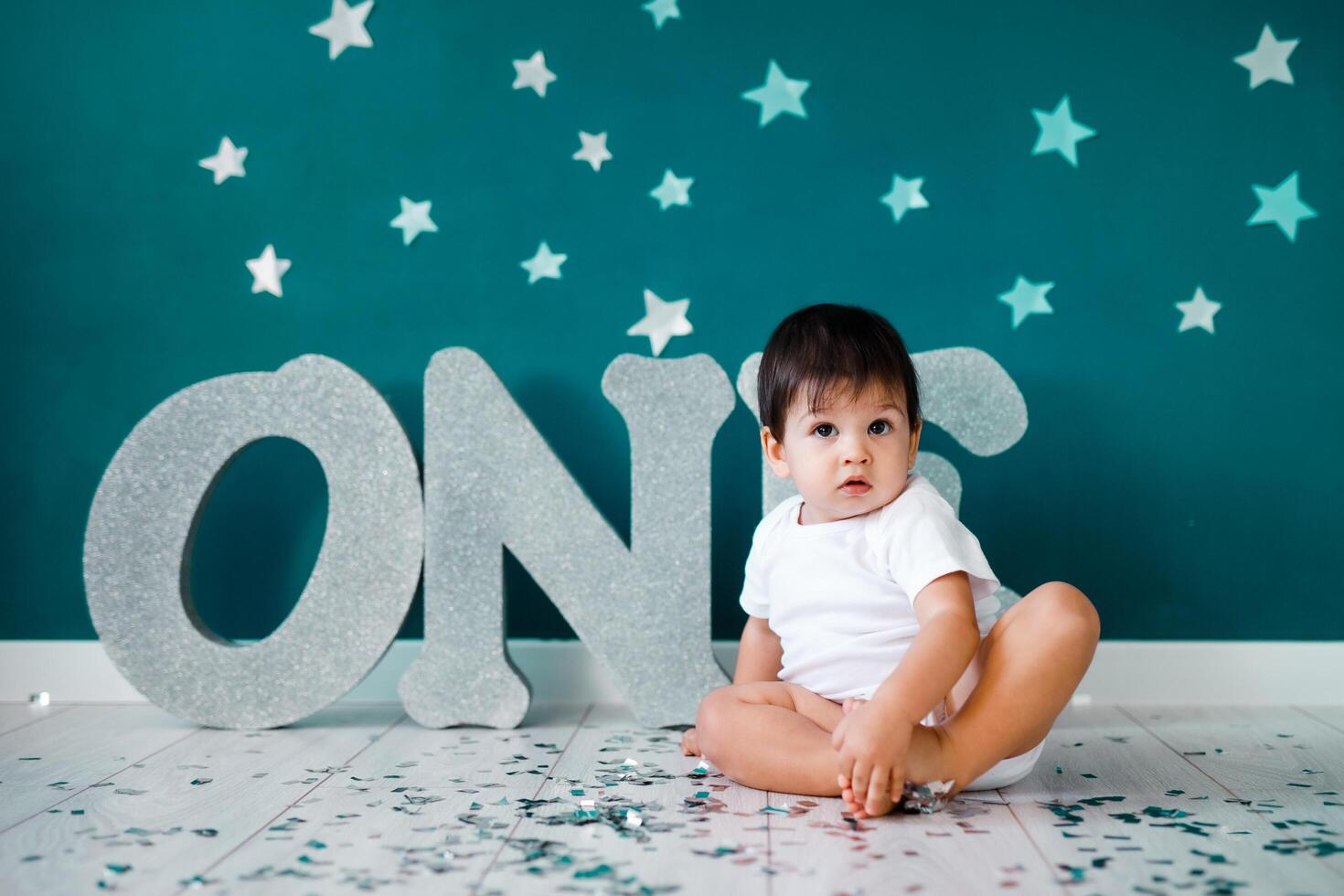 One year old baby boy in white t-shirt celebrates first birthday near silver letters ONE on green background with stars photo