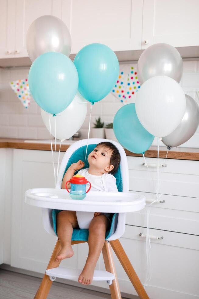 Little baby boy sitting in blue high chair at home on white kitchen and drinking water from sippy cup on background with balloons. photo