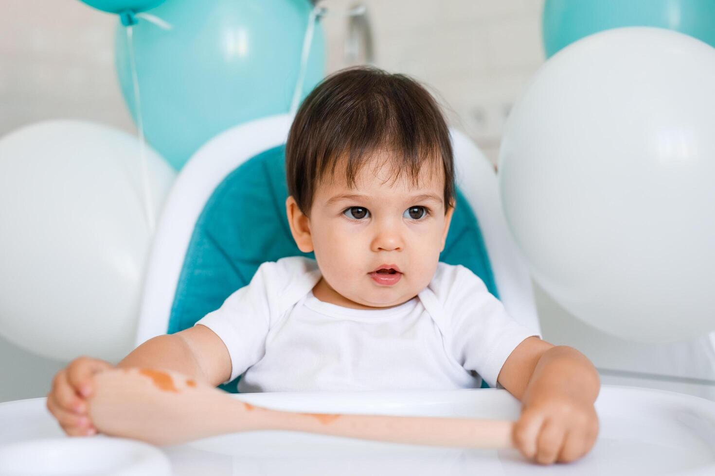 Little baby boy sitting in blue high chair at home on white kitchen and playing with wooden big spoon on background with balloons. photo