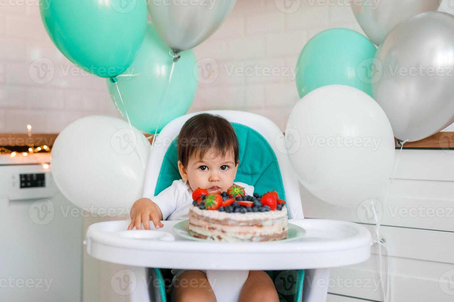 Little baby boy sitting in high chair in white kitchen and tasting first year cake with fruits on background with balloons. photo