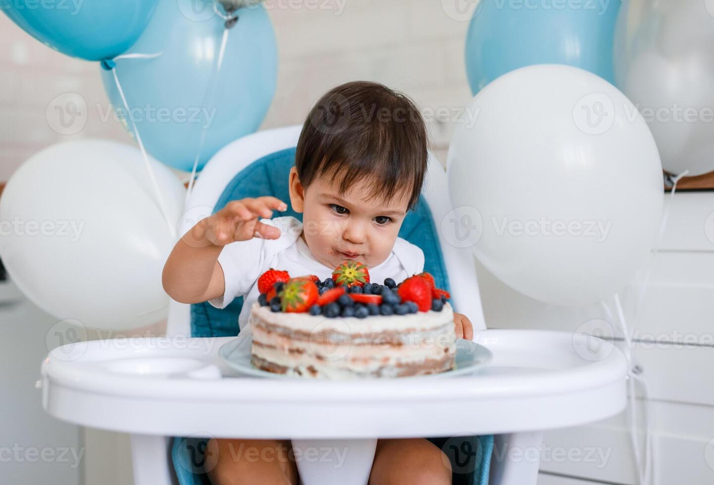 Little baby boy sitting in high chair in white kitchen and tasting first year cake with fruits on background with balloons. photo