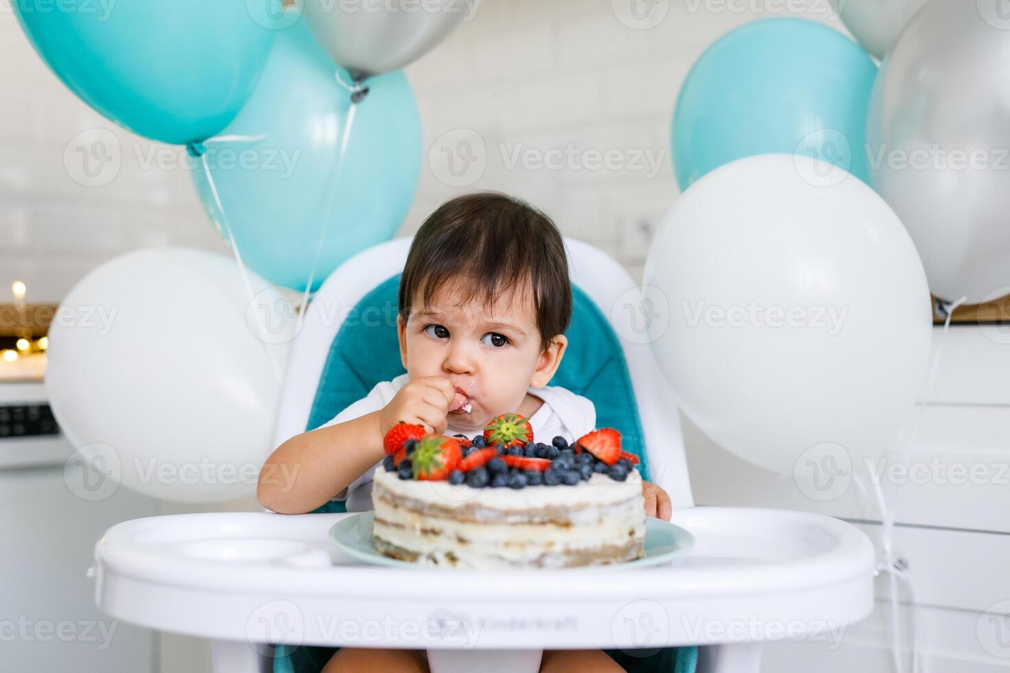 A one-year-old boy sits in a high chair in the kitchen eating birthday cake photo