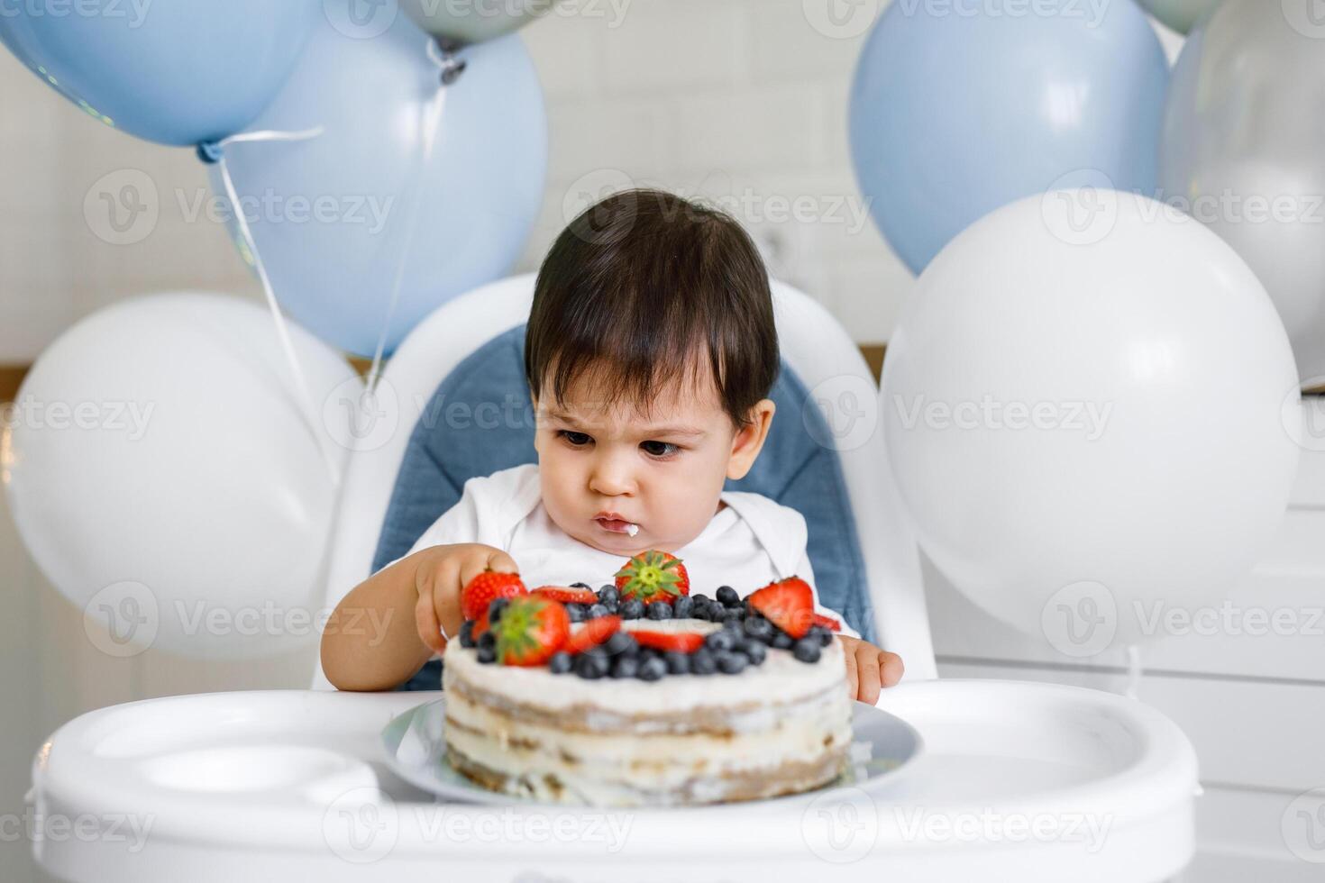 Little baby boy sitting in high chair in white kitchen and tasting first year cake with fruits on background with balloons. photo