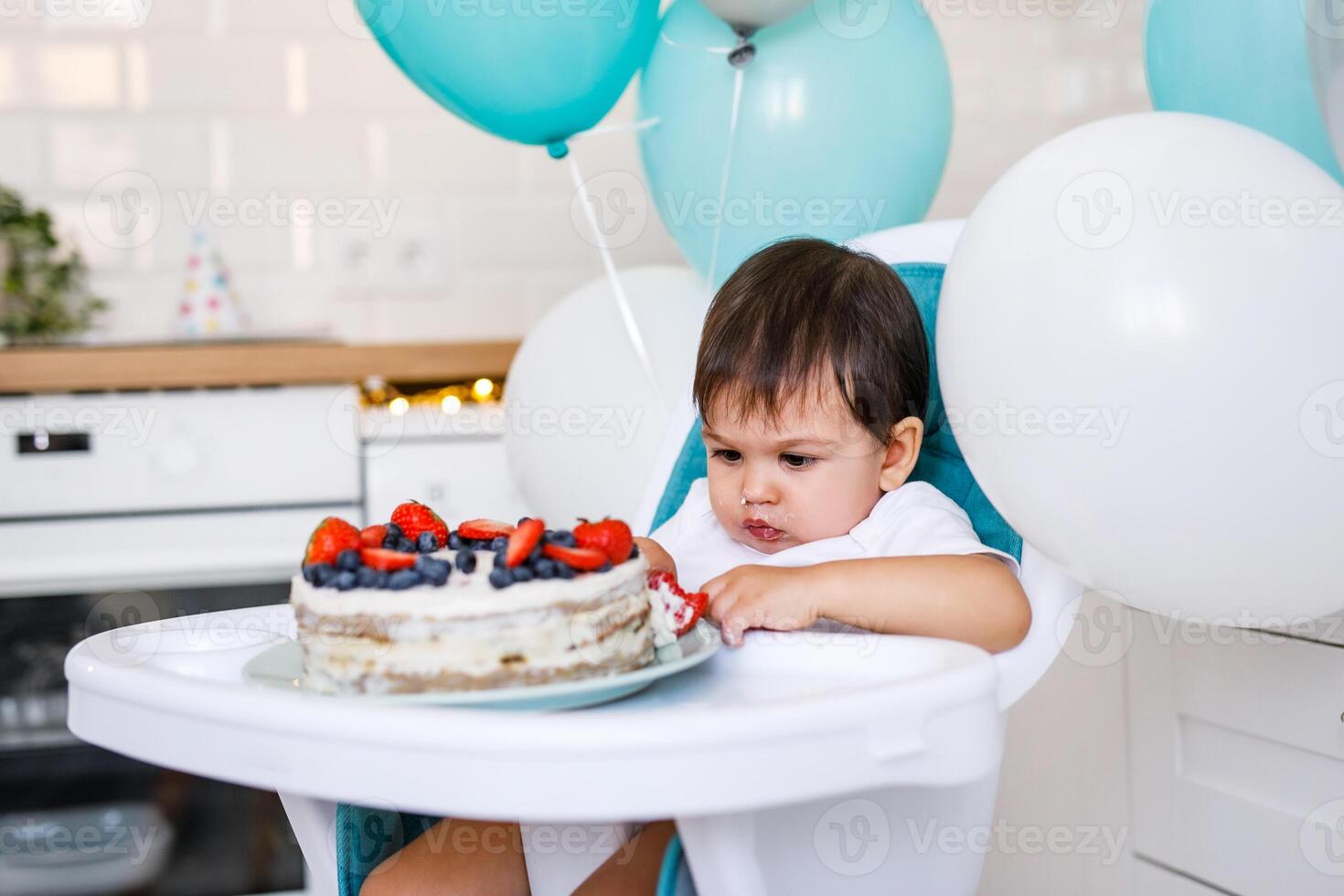 Little baby boy sitting in high chair in white kitchen and tasting first year cake with fruits on background with balloons. photo
