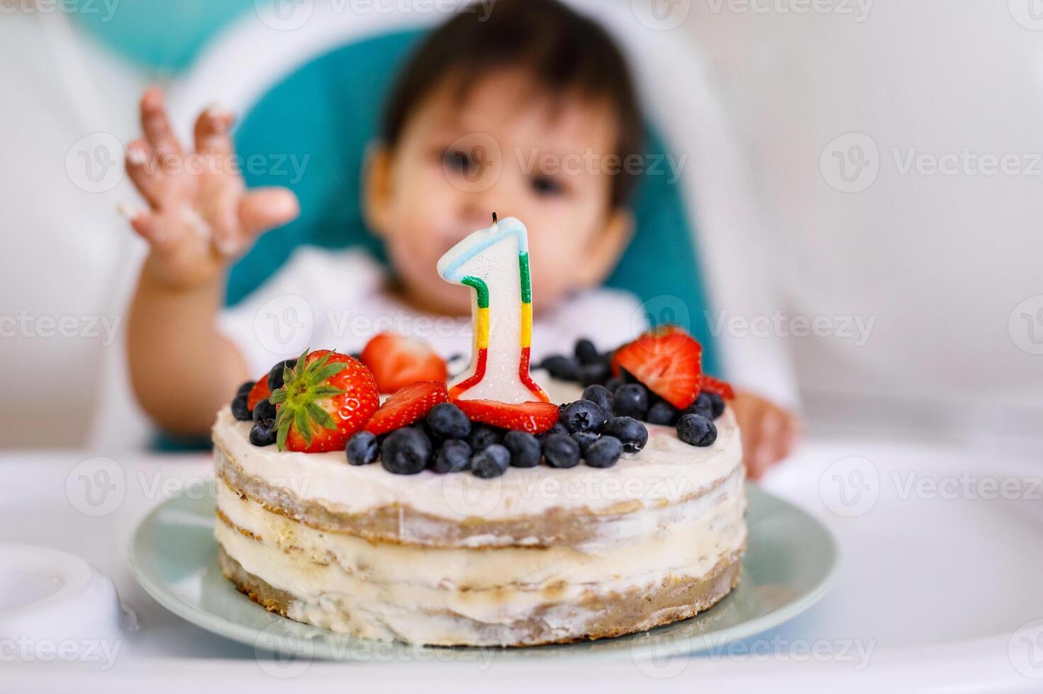 Little baby boy sitting in high chair in white kitchen and tasting first year cake with fruits on background with balloons. photo