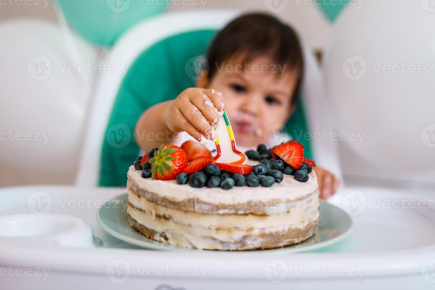 Little baby boy sitting in high chair in white kitchen and tasting first year cake with fruits on background with balloons. photo