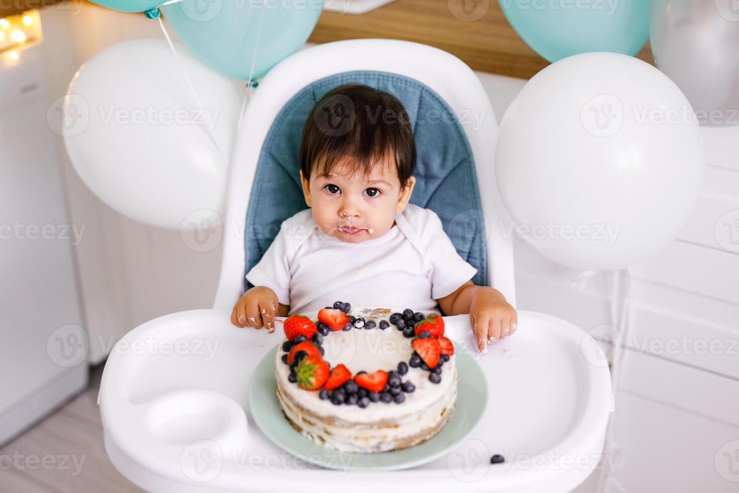 Little baby boy sitting in high chair in white kitchen and tasting first year cake with fruits on background with balloons. photo