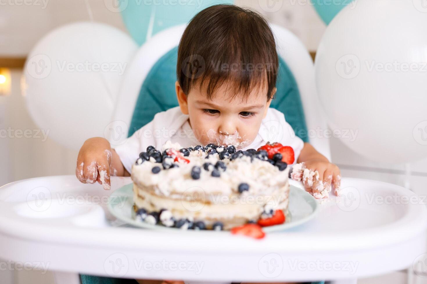 Little baby boy sitting in high chair in white kitchen and tasting first year cake with fruits on background with balloons. photo
