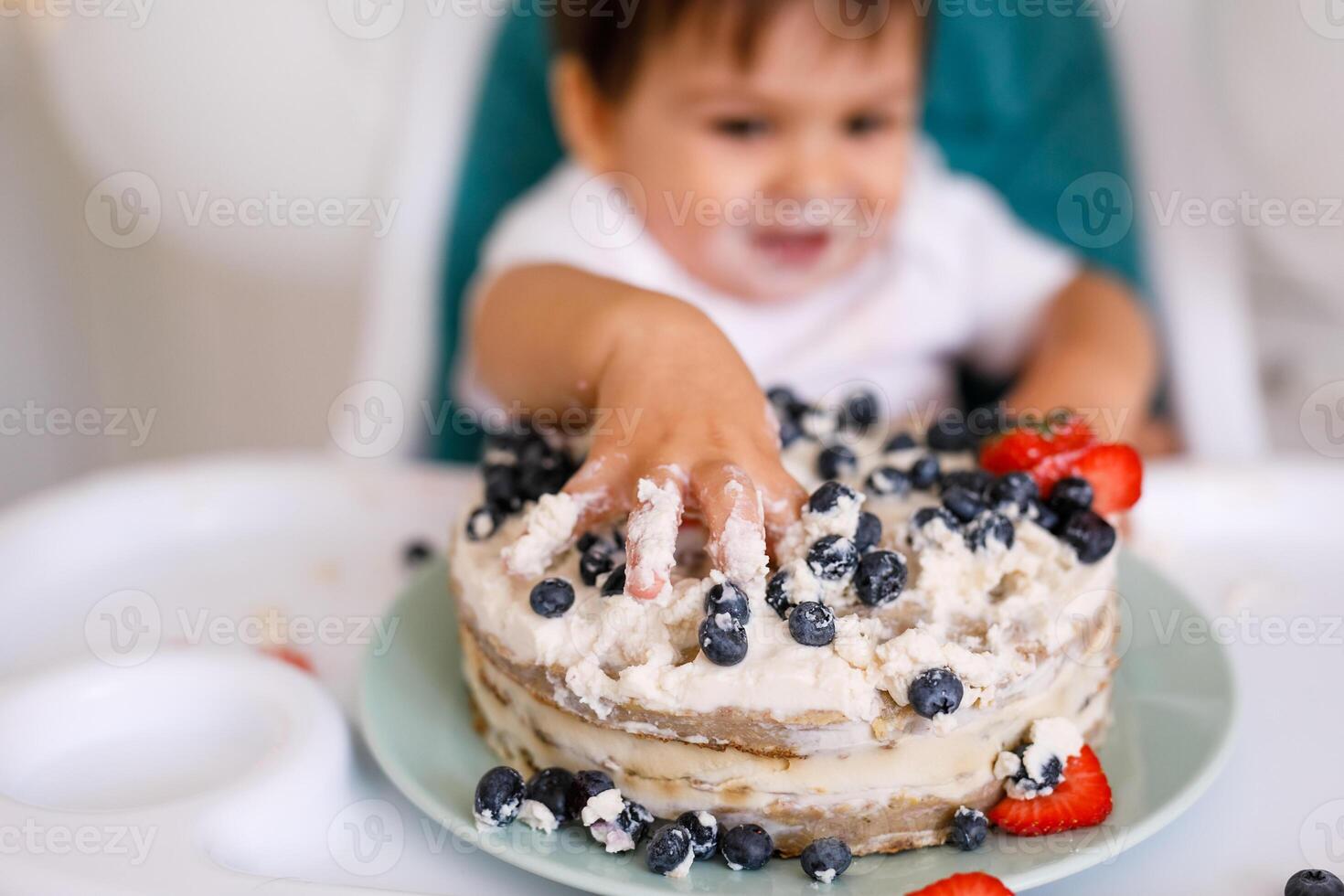 Little baby boy sitting in high chair in white kitchen and tasting first year cake with fruits on background with balloons. photo
