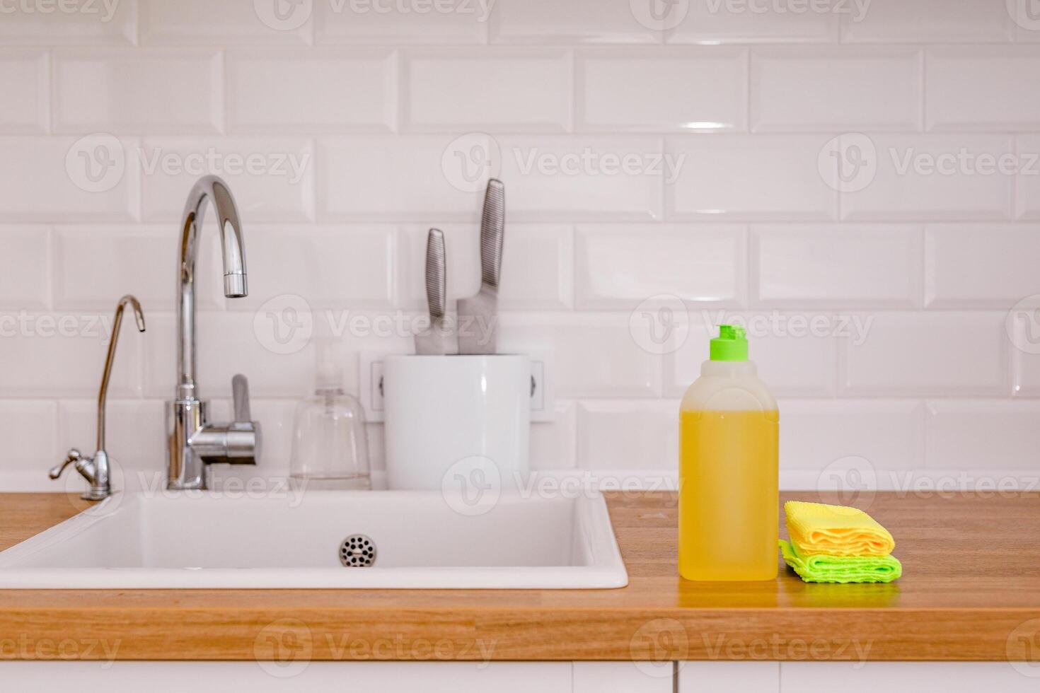 White plates, glass cups and forks in a clean white sink and two bottles of detergent and microfiber rags on a wooden table in a white kitchen photo