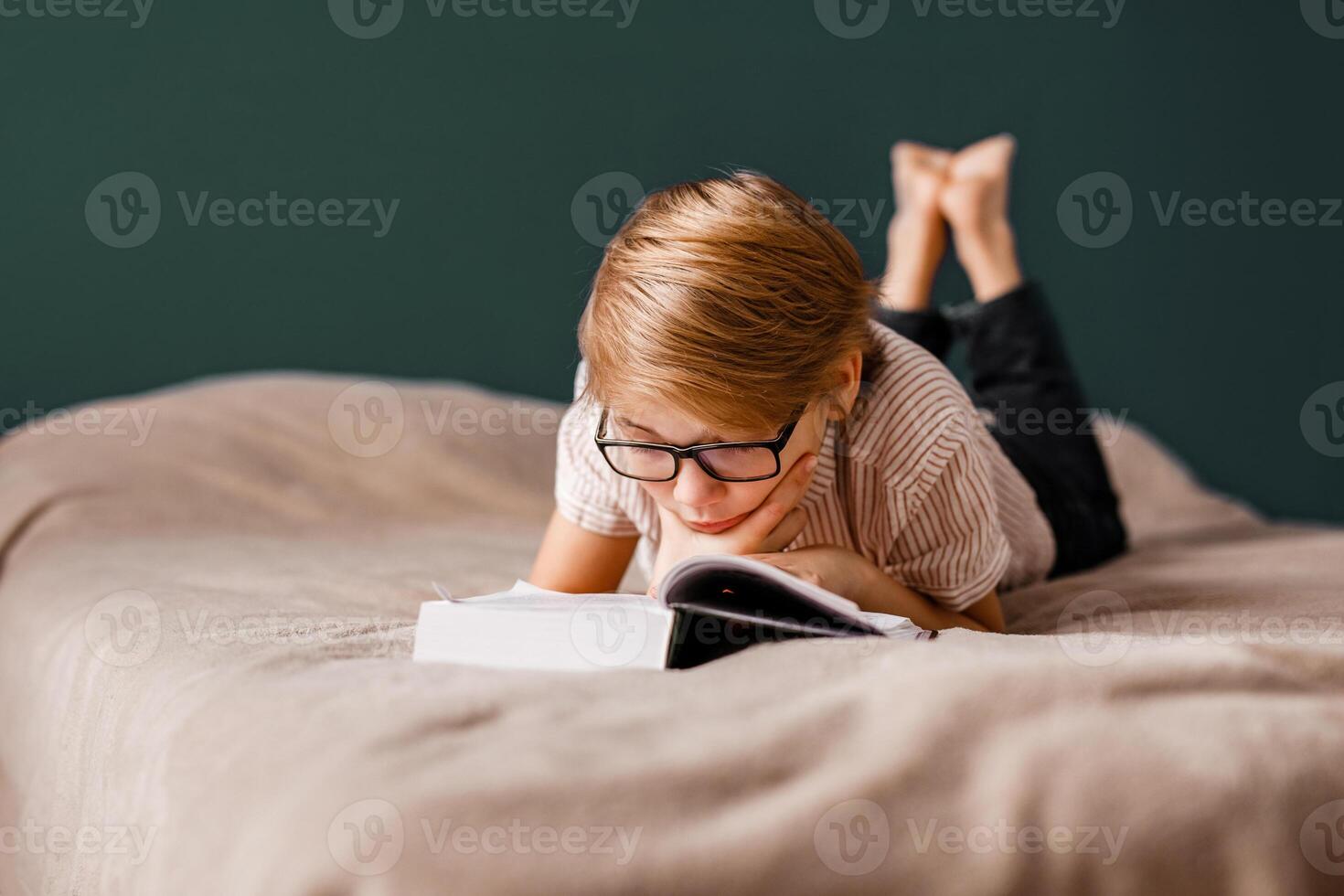 A 10-year-old boy with glasses is lying on the bed reading a big book photo