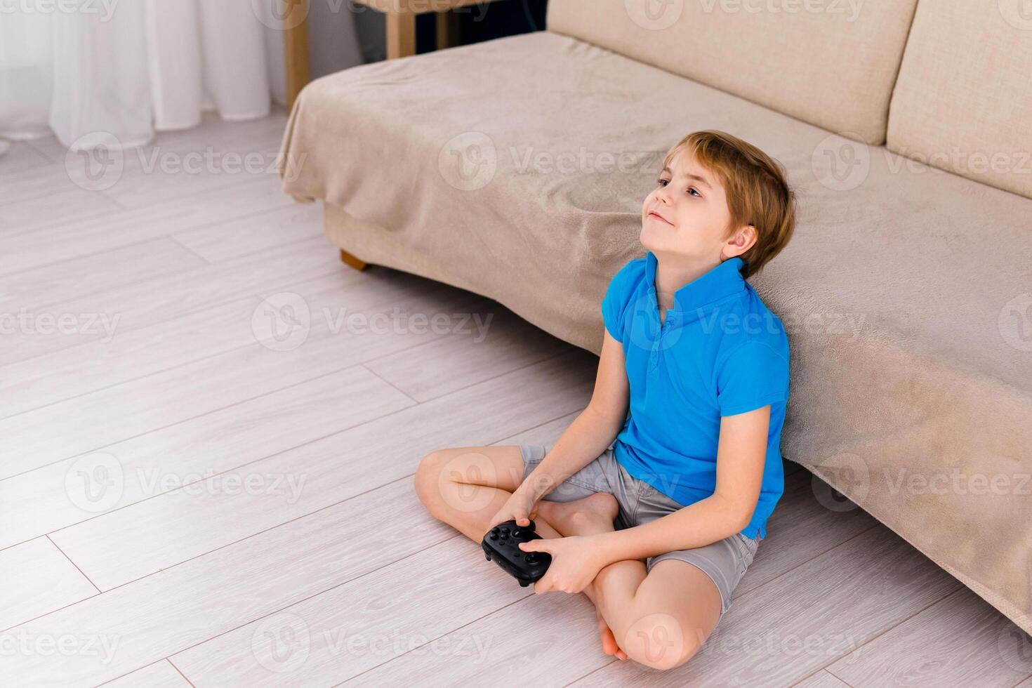 a 9-year-old boy in a blue T-shirt and big glasses sitting on the couch and playing at home with a gamepad. Blue background and free space for text photo