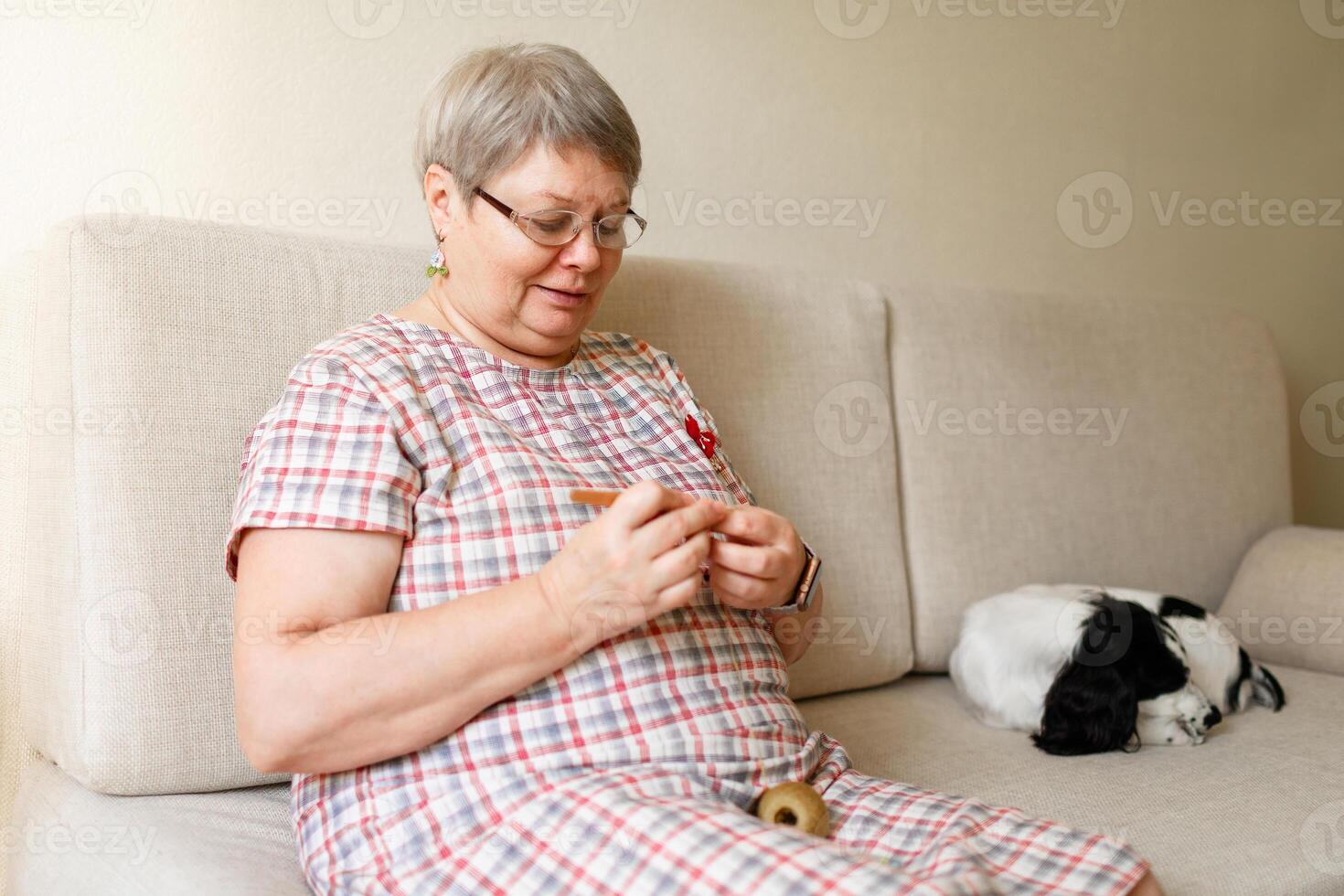 Elderly woman in glasses crocheting at home, a white dog sits beside her photo