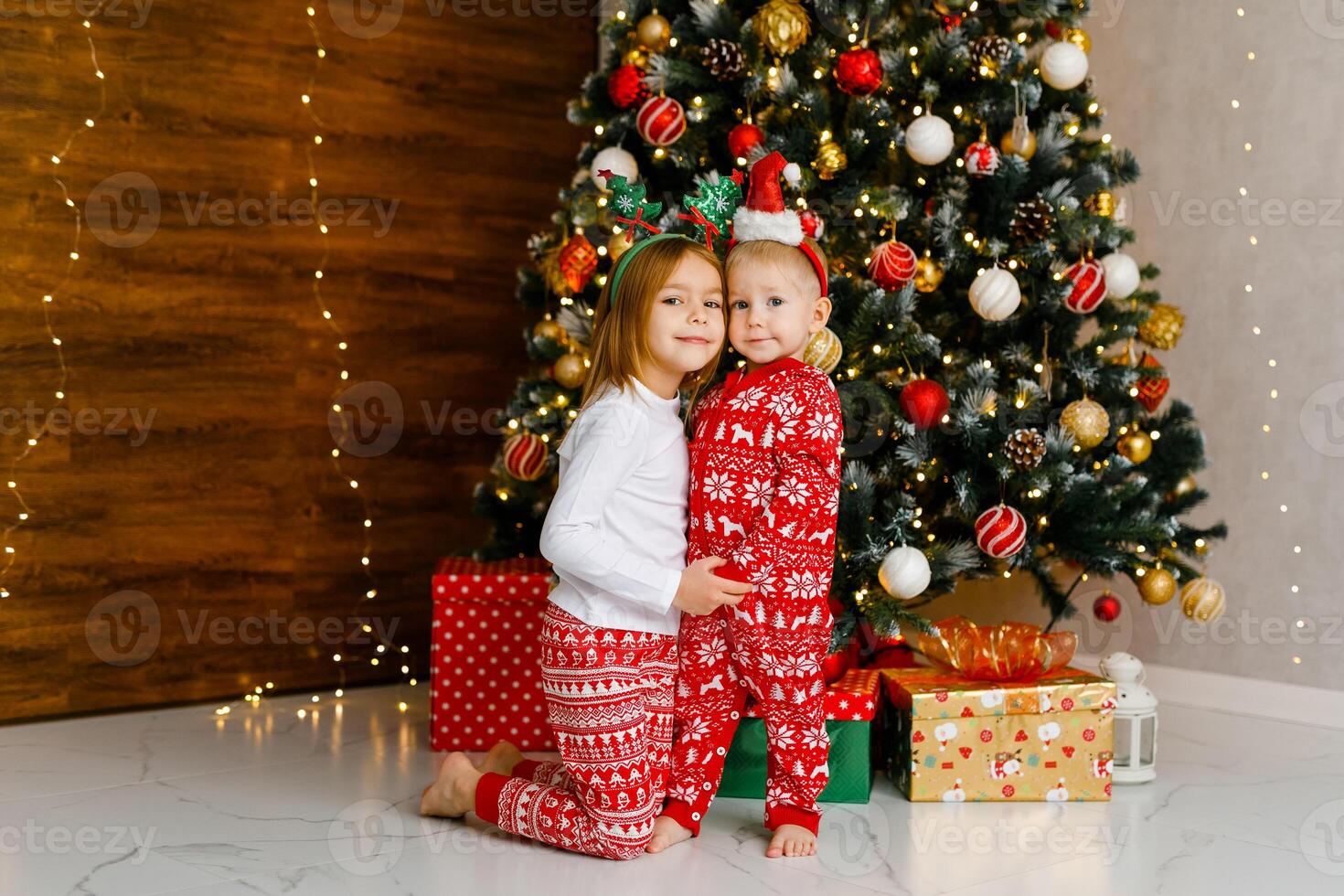 Little brother and sister in red pajamas are standing next to a Christmas tree photo