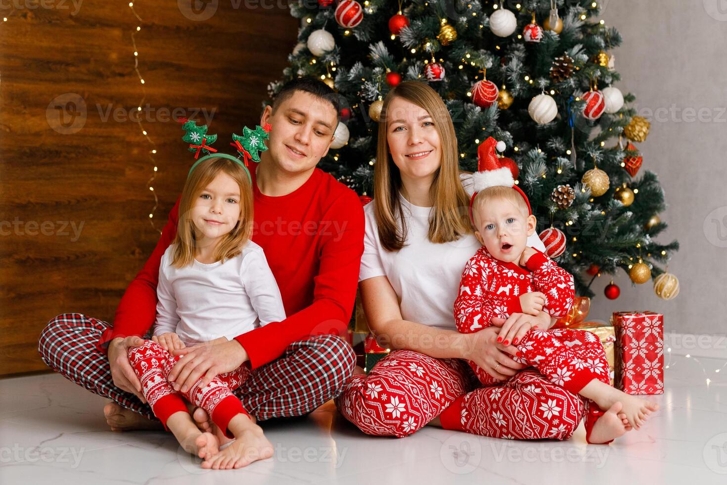Mom, Dad, and the brother and sister in red pajamas are playing and hugging in front of the Christmas tree photo