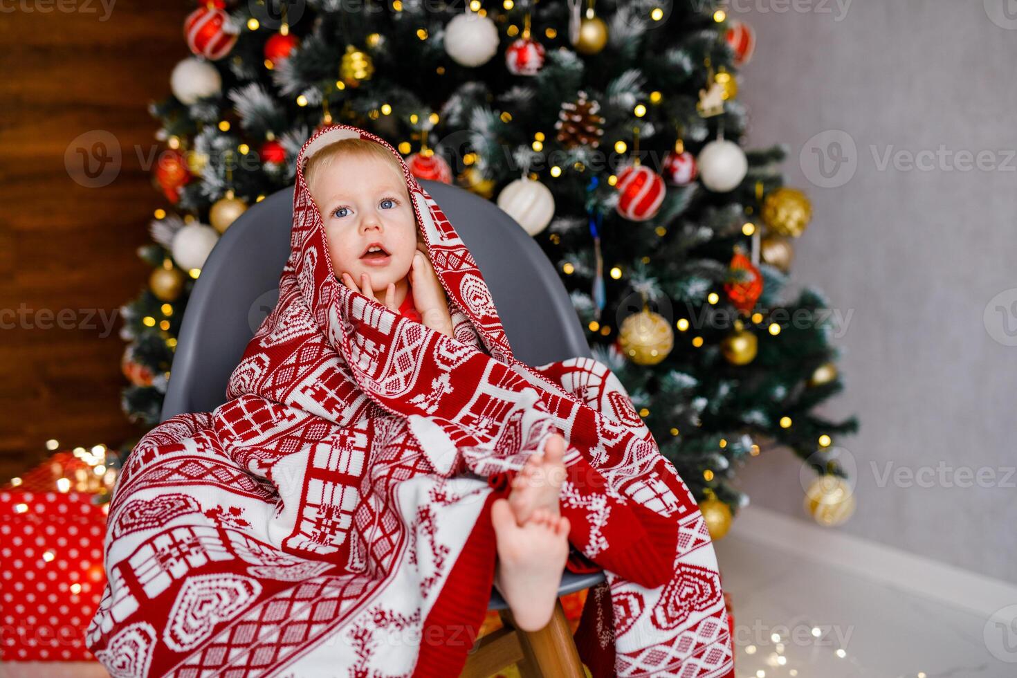 A 2-year-old boy is sitting in a chair wrapped in a warm blanket against the backdrop of a large Christmas tree photo