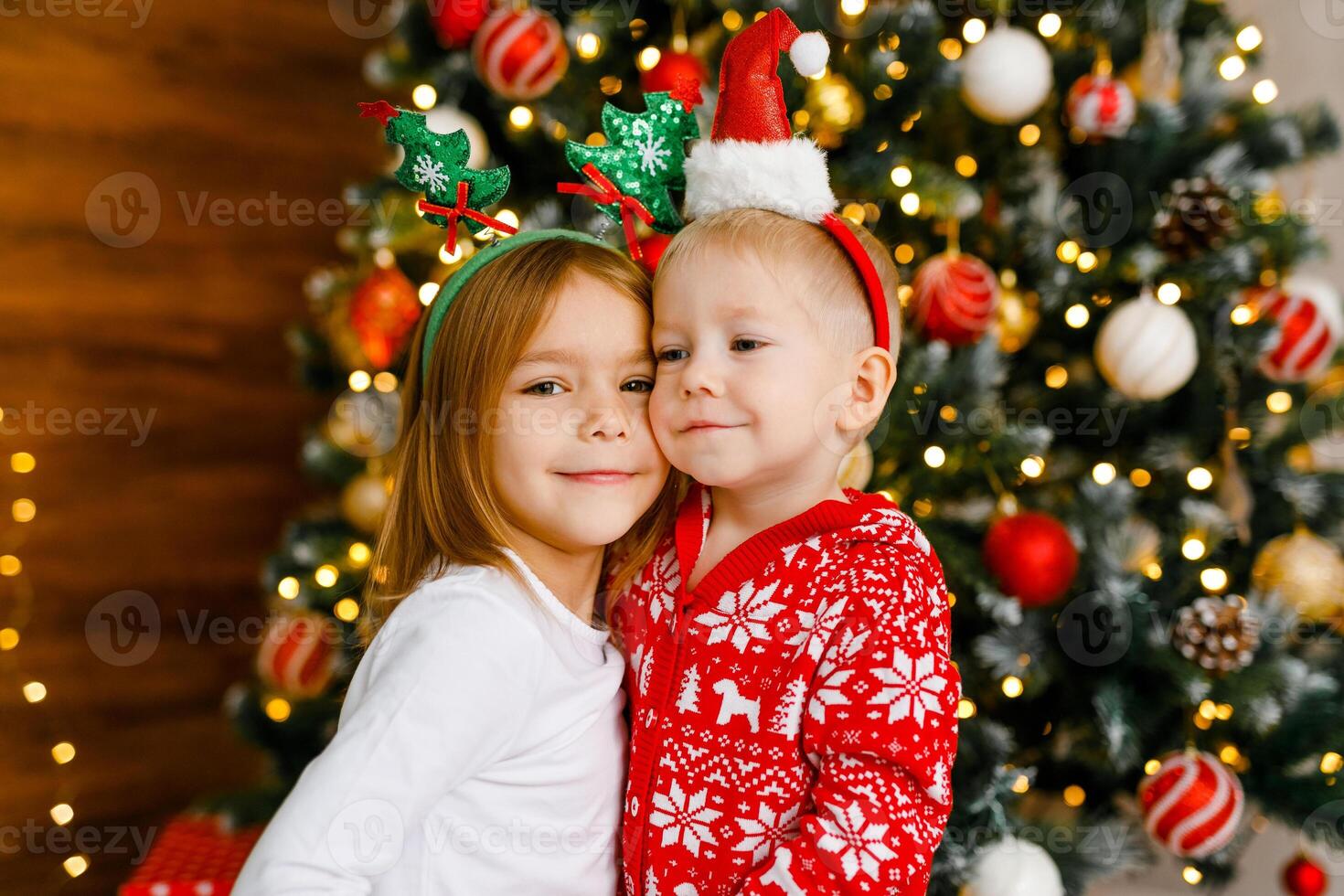 Young boy and girl in crimson and white nightwear with playful holiday headbands are standing beside the Christmas tree. photo