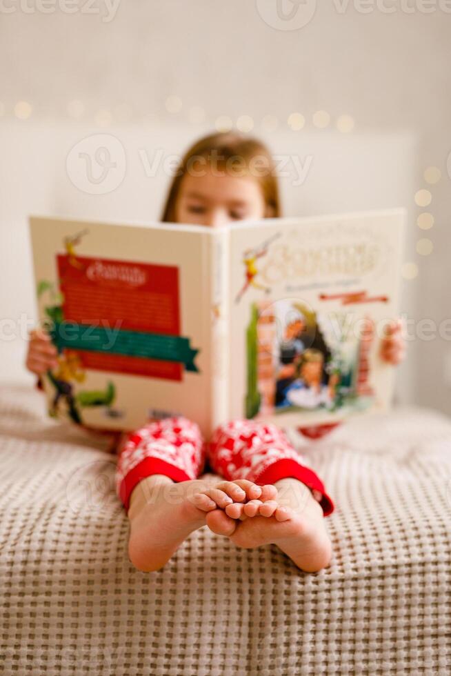 A girl in a Christmas pajama is reading a large book on the bed, focusing on her feet photo