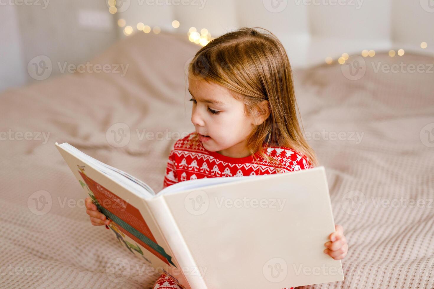 A little girl wearing holiday sleepwear is on the bed, reading a big book photo