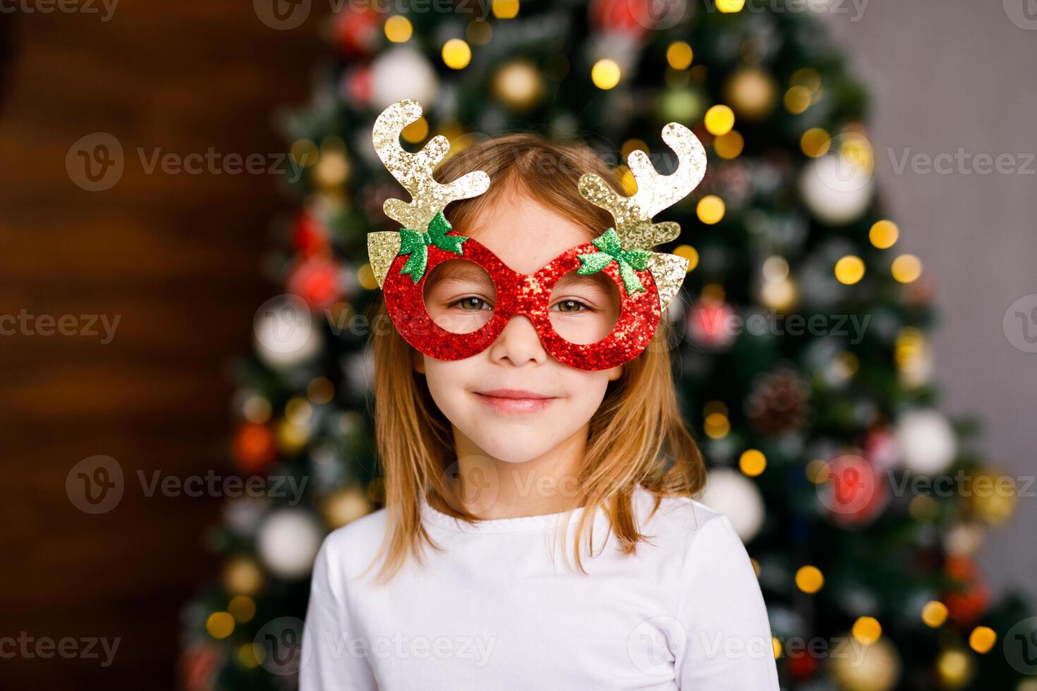 A six-year-old girl in shiny red glasses with golden horns is posing against the background of a Christmas tree photo