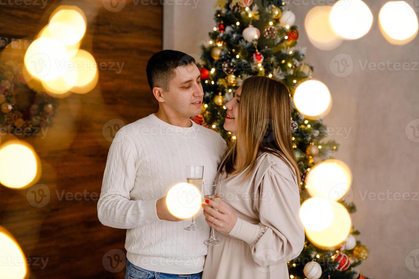A couple in love hugs and sips champagne in front of Christmas tree with bokeh from twinkling garland lights all around photo