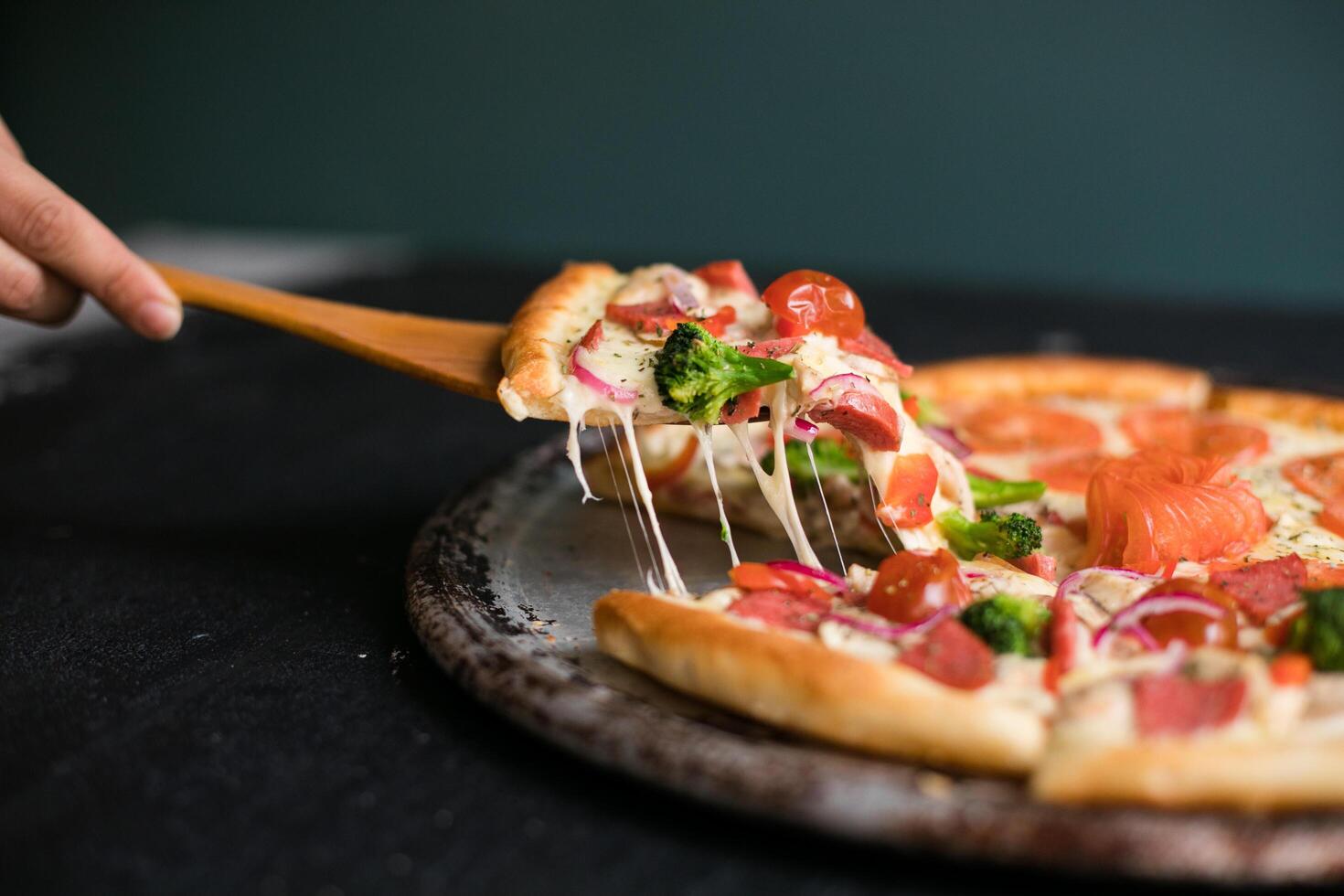 Delicious cheese stringy slice lifted of full supreme pizza baked fresh out of the oven next to ingredients photo